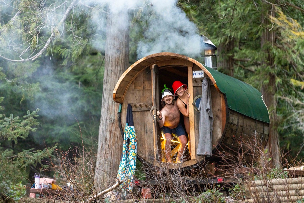 Two people wearing felt sauna hats in the doorway of a steaming wood-fired barrel sauna
